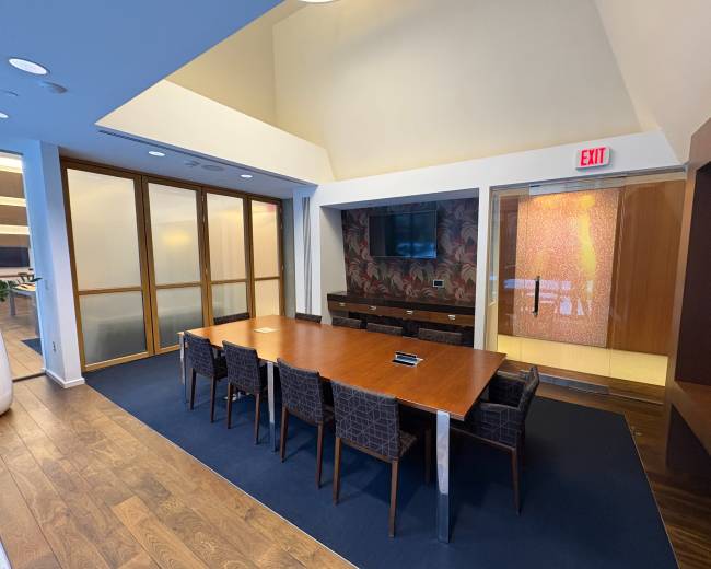 The image shows a modern conference room featuring a large wooden table surrounded by dark upholstered chairs, with a wall-mounted screen and glass doors leading to another space.