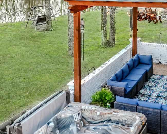 A hot tub sits on a patio beside a seating area and a view of a reflecting pond and trees.