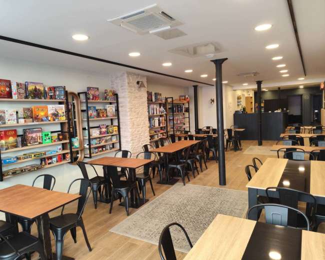The image shows a modern game café with neatly arranged wooden tables, chairs, and shelves filled with various board games.