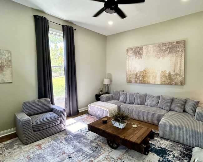 The image shows a modern living room with a sectional sofa, an accent chair, a wooden coffee table, and a large abstract painting on the wall.