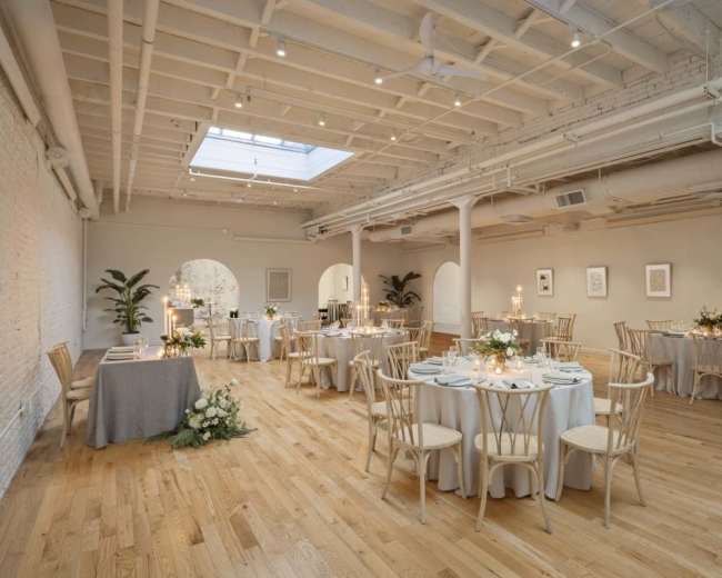 The image shows a beautifully arranged event space with round tables set for dining, soft lighting, and plants in each corner.