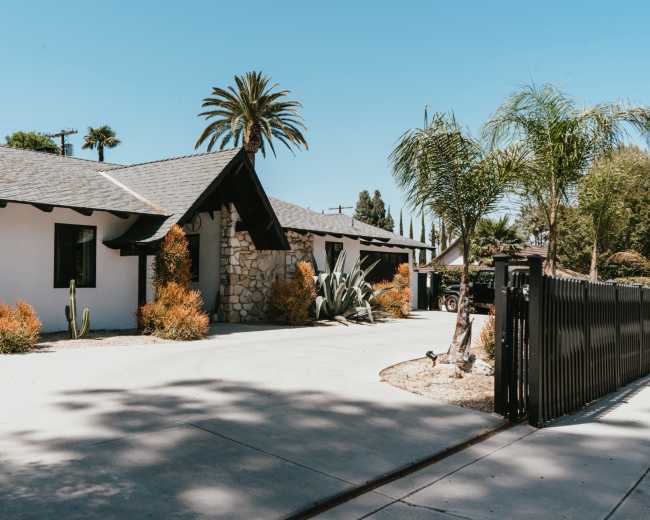 The image shows a single-story house with a stone facade, surrounded by palm trees and shrubs, set against a clear blue sky.