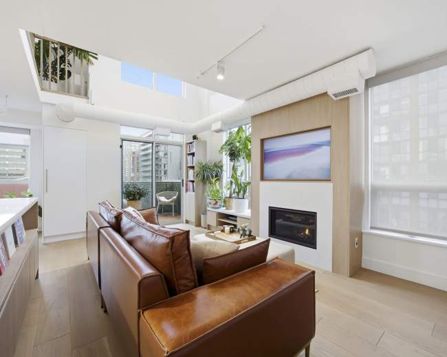 The image shows a bright, modern living room with a leather sofa, a fireplace, and large windows that provide natural light and a view of a balcony.