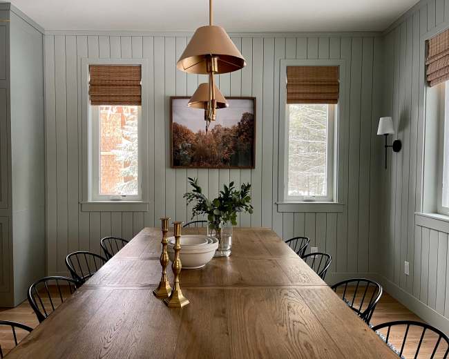 A spacious dining room features a long wooden table surrounded by black chairs, with a large pendant light overhead and wall art displayed between two windows.