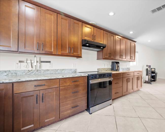 The image shows a modern kitchen with wooden cabinets, a black stove, and granite countertops, featuring a light-colored tile floor.