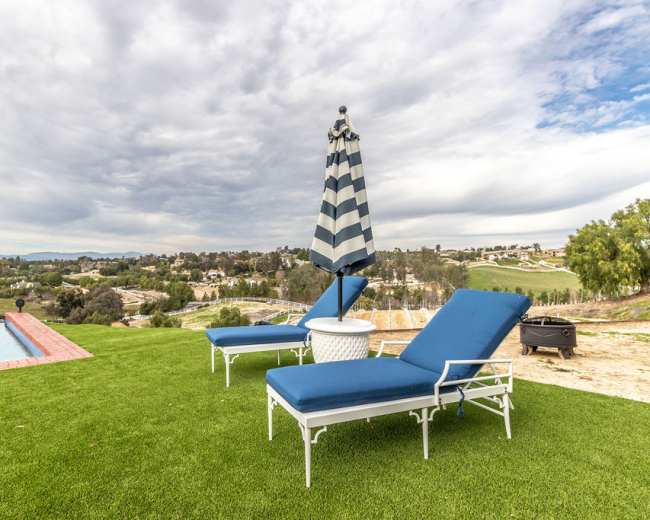 Two blue lounge chairs and a striped umbrella sit on a green lawn overlooking a valley with homes and rolling hills in the background under a cloudy sky.