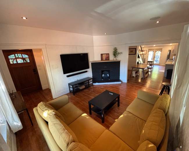 The image shows a living room with a yellow sectional sofa, a black coffee table, a wall-mounted TV, and a fireplace, adjacent to an entryway and a dining area.
