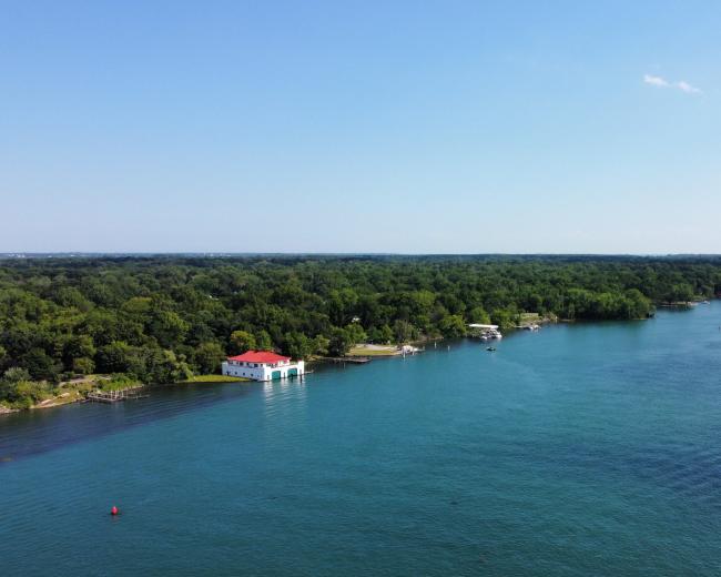 The image shows a calm river flowing alongside a forested area, with a white building featuring a red roof on the riverbank.