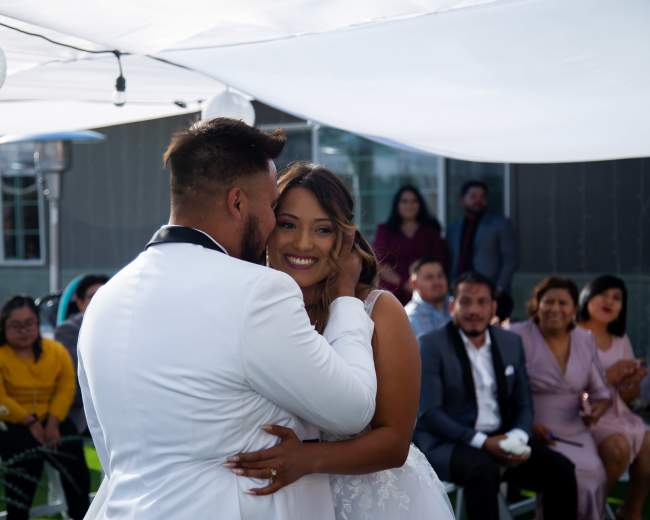 A couple dances closely under a white tent while guests watch in a garden setting.