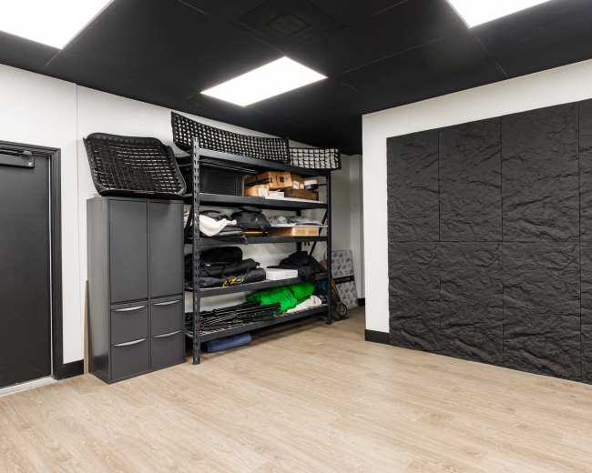 The image shows a storage room with shelving units filled with various items and a black wall panel.