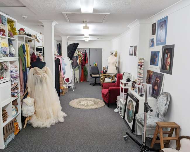 A spacious dressmaking studio featuring a variety of colorful fabrics, a mannequin dressed in a formal gown, and numerous photos and props arranged throughout the room.