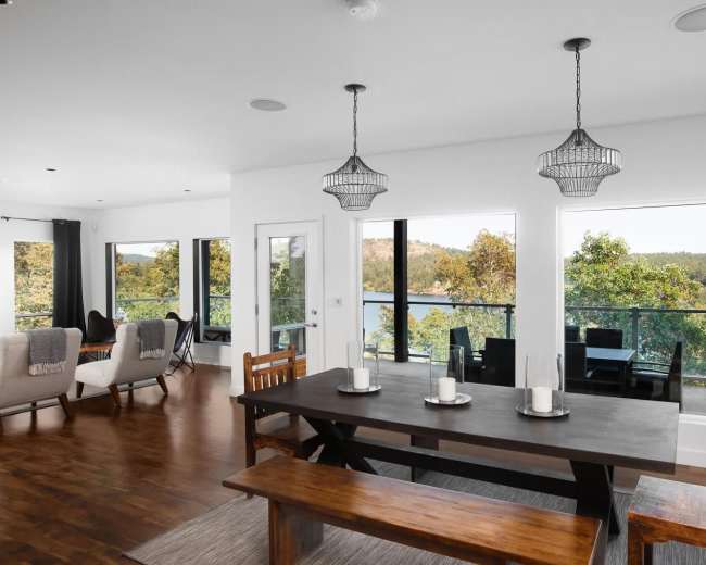 A dining area features a large table with a bench and chairs, surrounded by windows overlooking a scenic view.