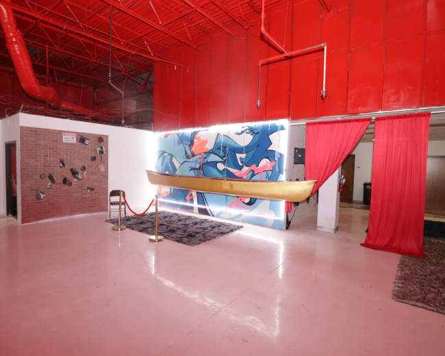 An interior space with a bright red ceiling, a mural of blue figures on the wall, a suspended canoe, and red curtains framing the area.