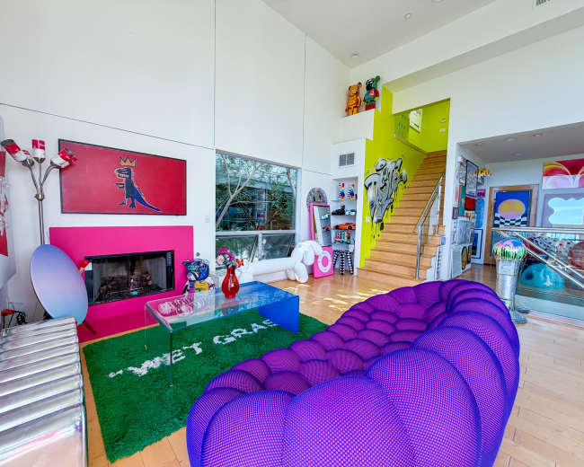 The image shows a spacious living room with a purple couch, a modern coffee table, and colorful artwork on the walls, alongside a staircase leading to an upper level.