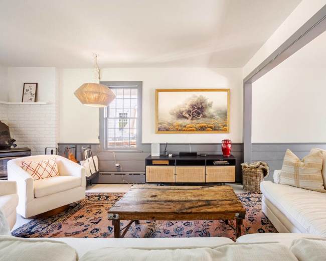 The image shows a cozy living room featuring two white sofas, a wooden coffee table, a large painting on the wall, and various decorative elements like a floor lamp and a woven basket.