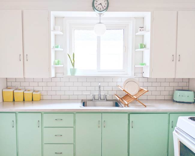 A bright kitchen featuring mint green cabinets, white countertops, and a window over the sink with dishes drying on a wooden rack.