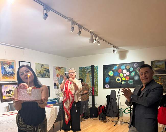 A woman in a black dress performs in front of an audience in an art gallery, while a man applauds and another person stands holding a scarf.