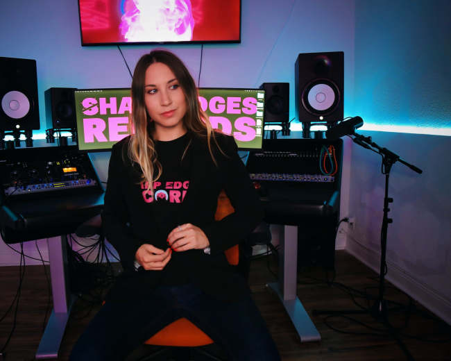 A woman sits in a chair in a music studio surrounded by audio equipment and monitors, with a colorful screen displaying "SHARP EDGES RECORDS" in the background.