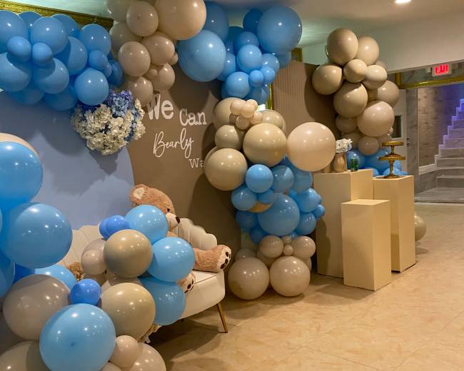 A decorated event space featuring a wall adorned with clusters of blue, beige, and white balloons, alongside a small seating area and decorative elements.