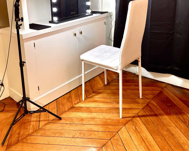 The image shows a makeup vanity with a lit mirror, a white chair, and shelving above it, all positioned against a dark curtain.