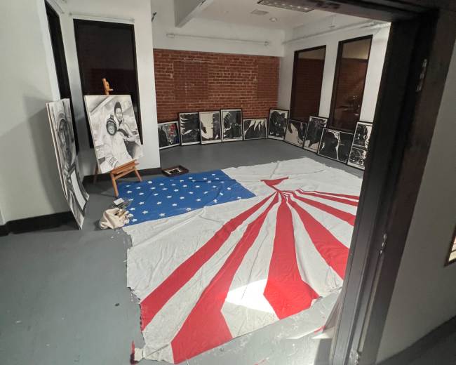 The image shows a room with a large red and white fabric arranged in a starburst pattern on the floor, surrounded by black and white framed photographs leaning against the walls.