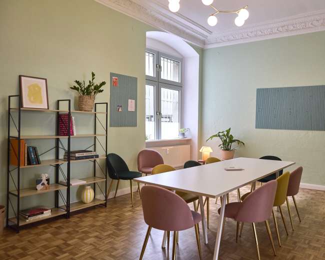 The image shows a light green room with a long white table surrounded by multi-colored chairs, a bookshelf with decorative items, and a window letting in natural light.
