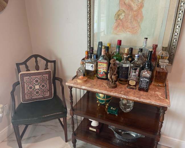 A wooden table topped with various bottles of liquor is positioned beside a chair and a framed artwork featuring two figures.