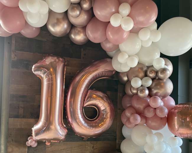 A decorative balloon arch with a large "16" balloon, incorporating shades of pink, white, and metallic colors against a wooden backdrop.