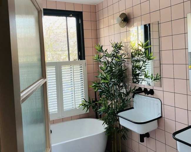 A bathroom with pink tiled walls, a freestanding tub, a small potted plant, and two mirrors above a wall-mounted sink.