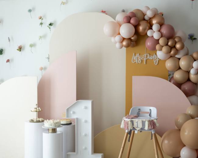 A decorated party space featuring a balloon arch, a cake display on white pedestals, and a high chair against a backdrop of soft pastel colors.