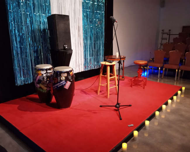A stage set up with two conga drums, a microphone on a stand, and two stools surrounded by candles, with chairs arranged in the background.