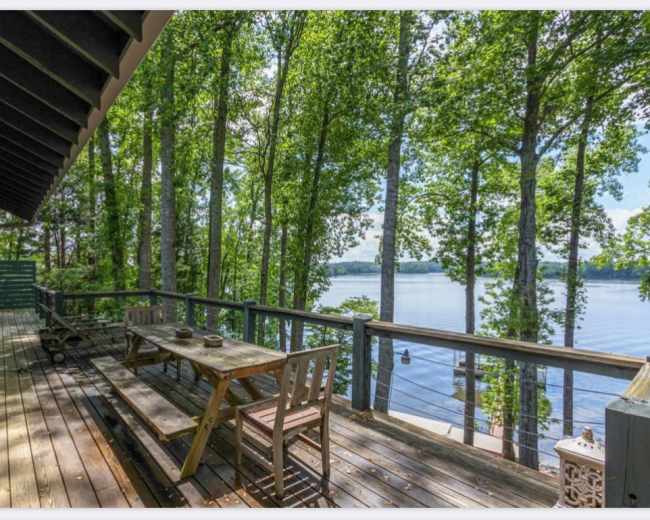The image shows a wooden deck overlooking a calm lake, surrounded by tall trees under a clear blue sky.