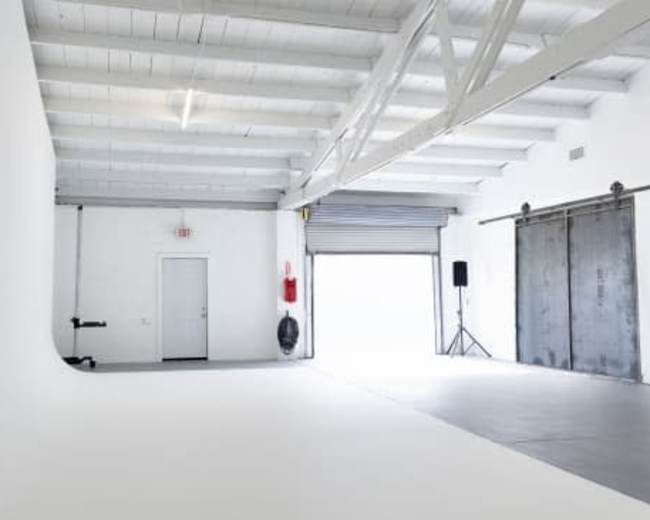 The image shows a spacious, minimalist photography studio with white walls, a smooth floor, and various equipment along one side.