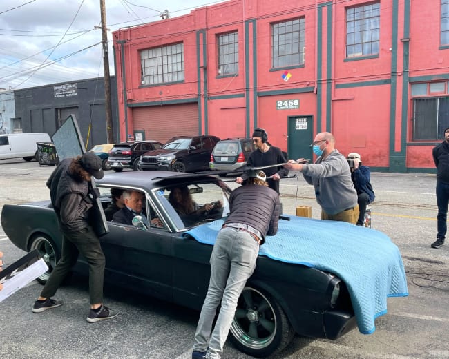 A group of people is filming a scene involving a black car in an urban setting with colorful buildings in the background.