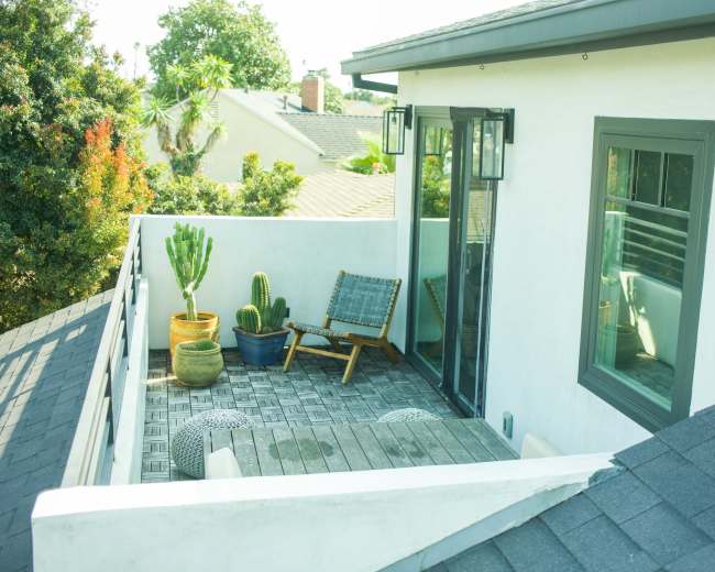 A terrace with a wooden floor, potted cacti, lounge chairs, and large windows, surrounded by greenery.