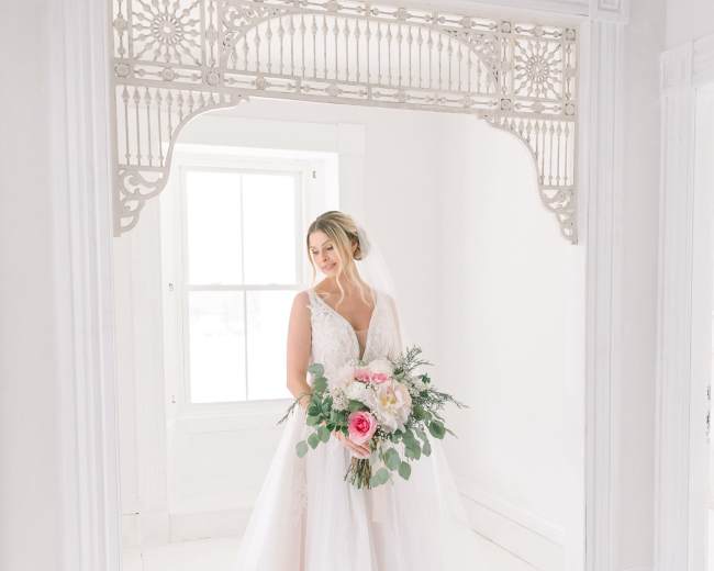 A bride holding a bouquet stands in a bright room adorned with ornate trim.