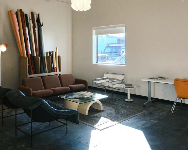 The image shows a modern, minimalist interior with a brown sofa, black chairs, a coffee table, and a window allowing natural light to illuminate the space.
