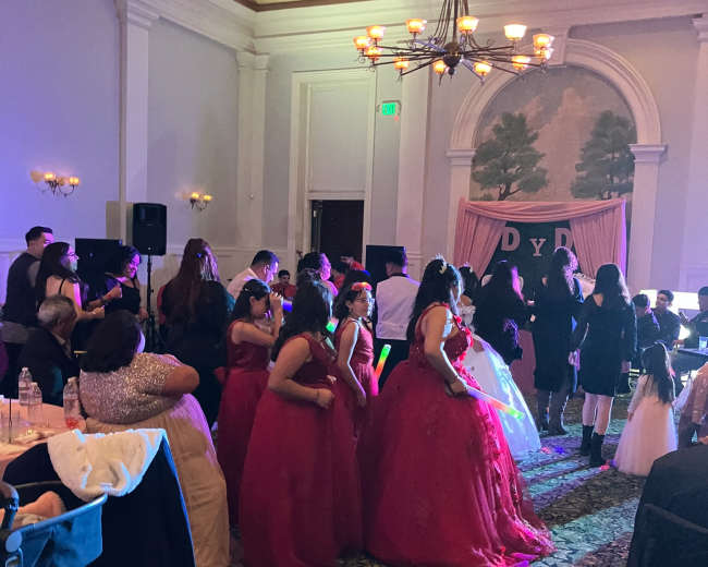 A group of people in formal attire, including several women in red dresses, are dancing during a festive event in a decorated indoor venue.