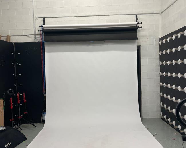A photography studio with a white backdrop set up against a textured wall, and various equipment arranged in the background.