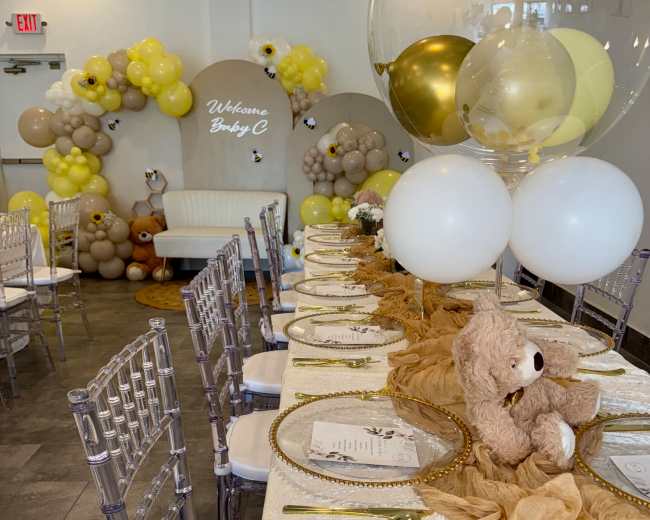 The image shows a decorated event space featuring a long table set with golden plates, a beige runner, teddy bear centerpieces, and colorful balloon arrangements in the background.