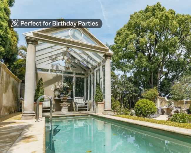 A glass-enclosed structure near a pool is surrounded by greenery and decorative plants.
