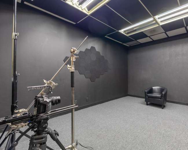 The image shows a dimly lit room with a black background, featuring a photography camera on a tripod and a single black chair in the corner.