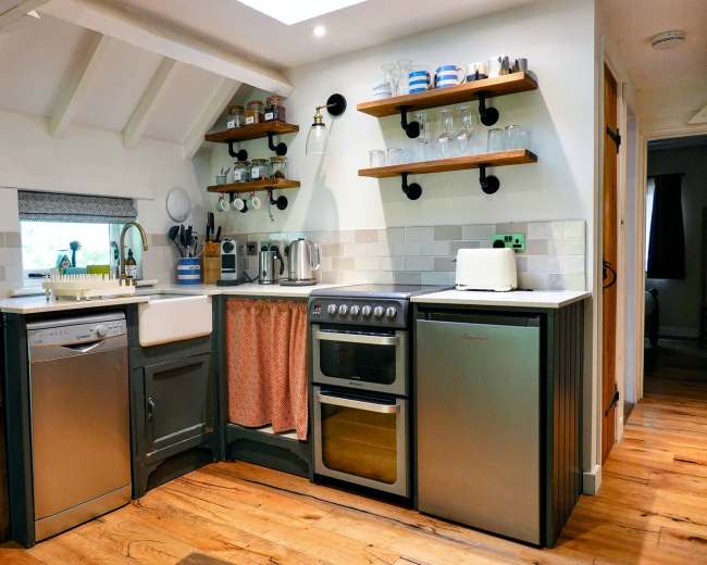 A compact kitchen features a sink, stove, dishwasher, and shelves with glassware and dishes, all arranged against a wooden floor and light-colored walls.
