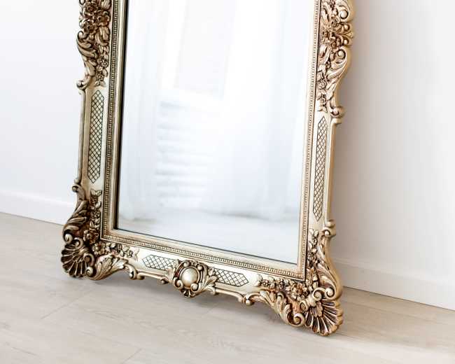 A large, ornate mirror with a decorative gold frame leans against a white wall on a light wooden floor.