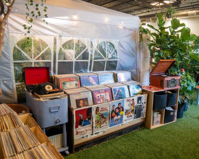 The image shows a collection of vinyl records displayed on shelves alongside a vintage record player, set in a green, plant-filled environment with a tent-like structure in the background.