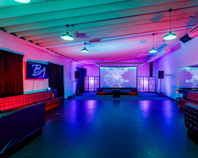 The image shows a dimly lit event space with colorful lighting, featuring a projection screen and seating areas along the walls.
