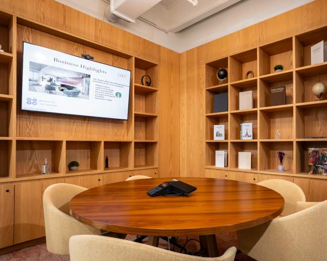 A conference room features a round wooden table surrounded by four upholstered chairs, with a large screen displaying business information on one wall and wooden shelves filled with books and decorative items on another.