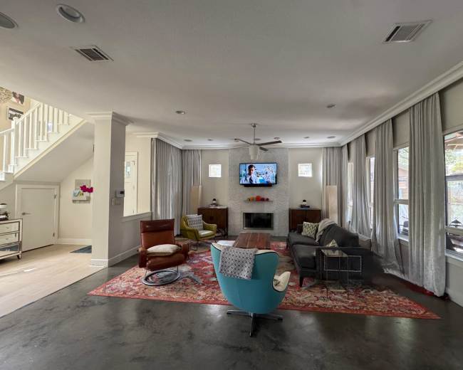 The image shows a modern living room with a television mounted on the wall, a mix of seating options including colorful chairs and a brown leather sofa, and large windows draped with light curtains.