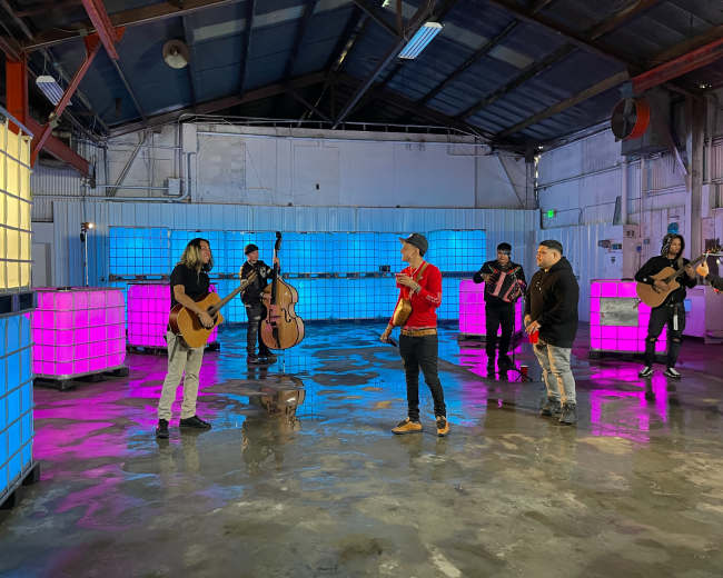 A group of musicians performs with various instruments in a warehouse illuminated by colorful lights.