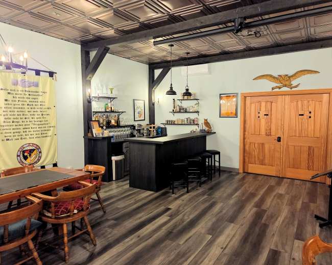 The image shows a cozy room with wooden flooring, a bar area with shelves, and two wooden doors at the far end.